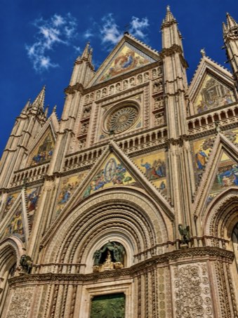 The west front of Orvieto cathedral seen from below &mdash; gold mosaics, a rose window and spires against a deep blue sky.