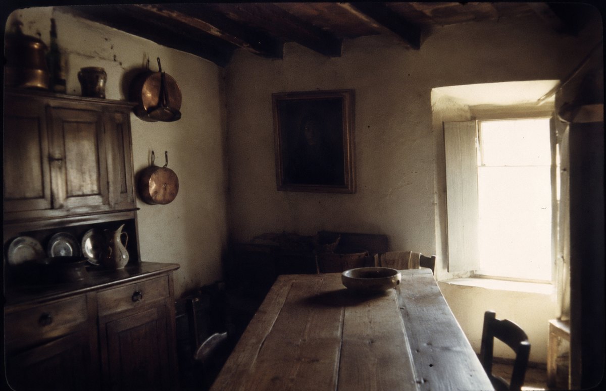 A murky old photograph of a farmhouse kitchen. On the back wall hangs a dark rectangle in a dull frame — a painting too dark to make out.